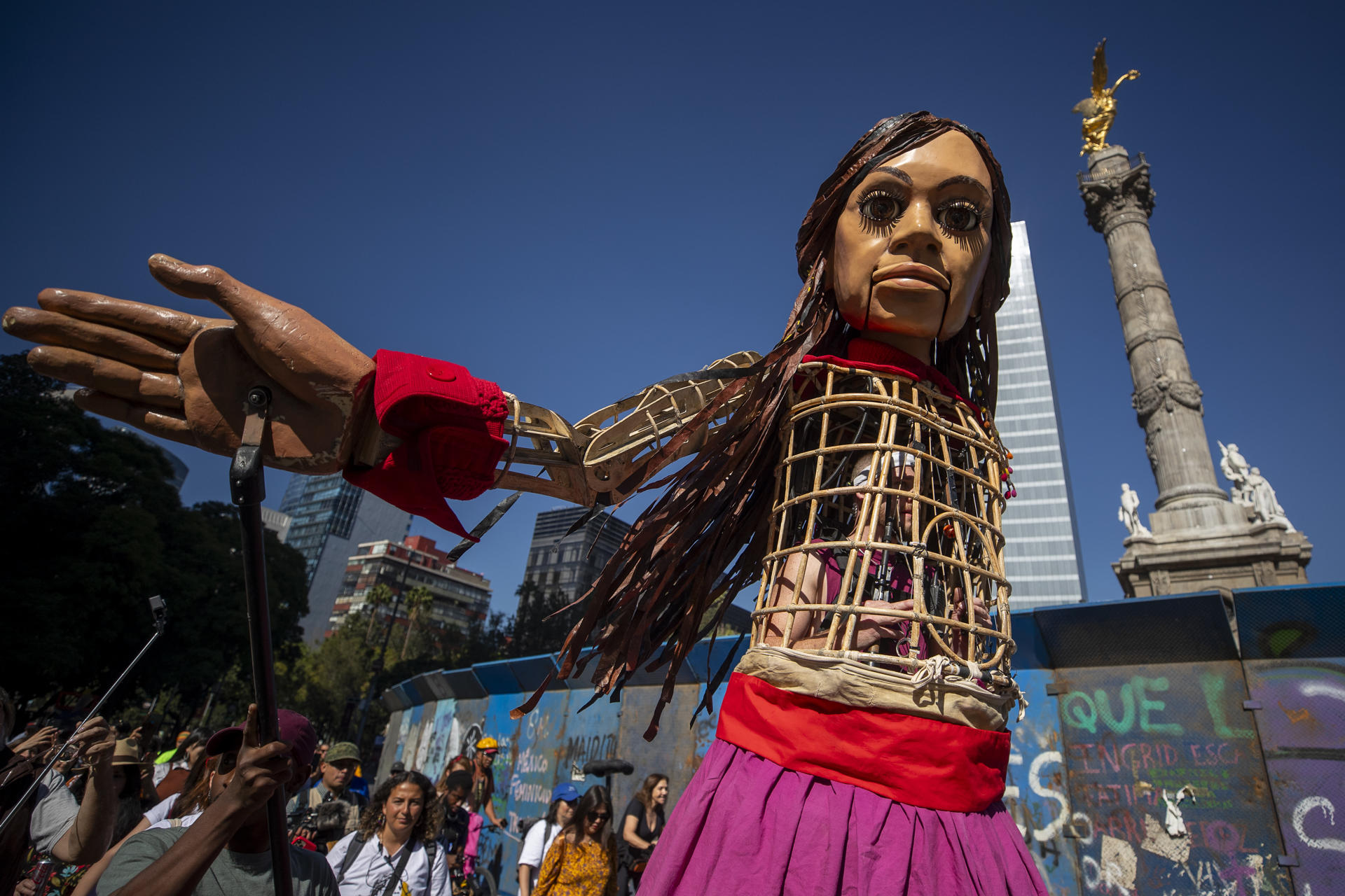 La marioneta Amal, que representa a una niña migrante siria, saluda a las personas durante su recorrido hoy, en la Glorieta de la Diana, a la Glorieta de los Desaparecidos, en avenida Paseo de la Reforma, en la Ciudad de México. EFE/ Isaac Esquivel

