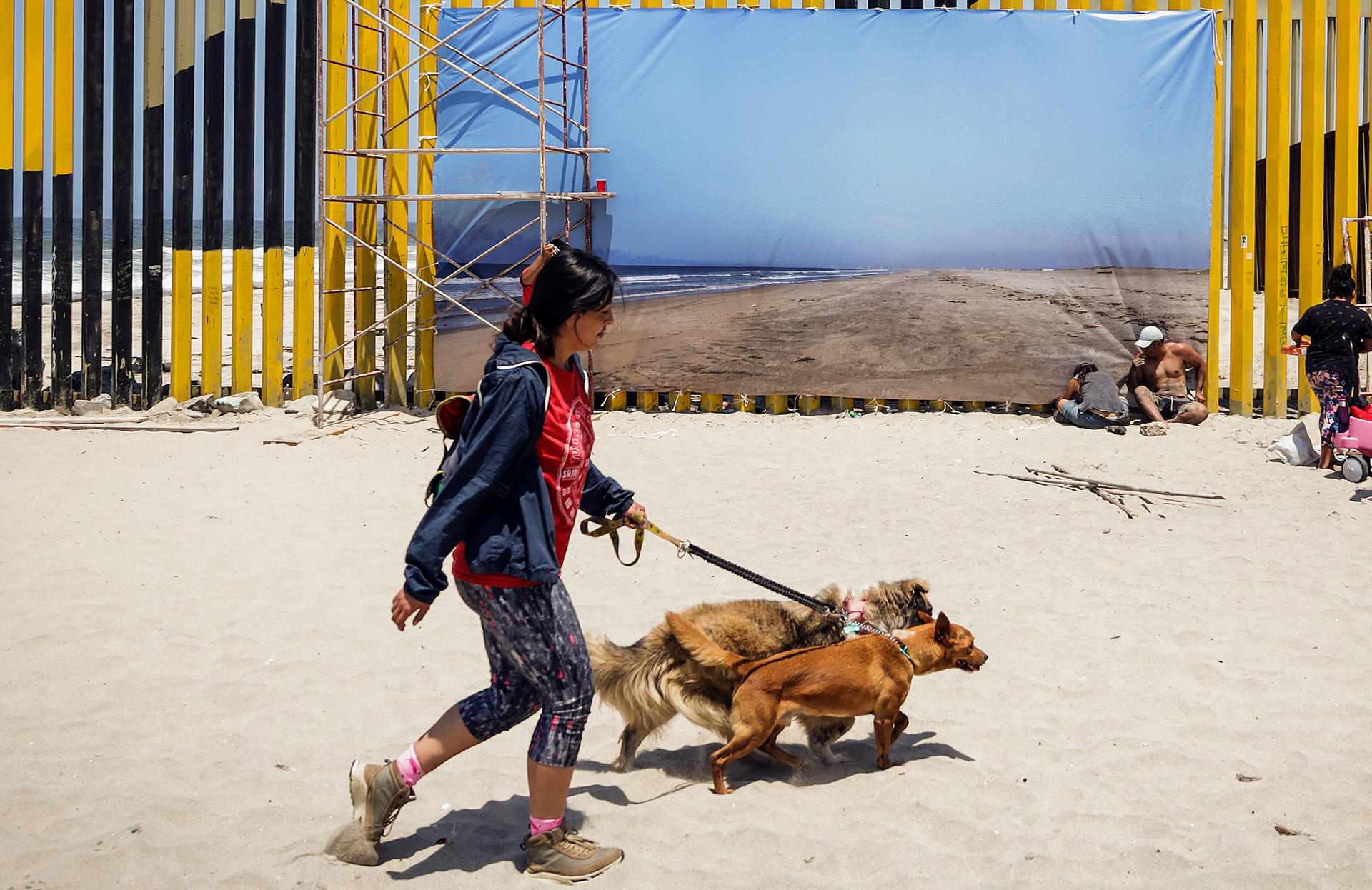 Una mujer camina con sus perros junto a una fotografía monumental colocada en el muro fronterizo, el 26 de julio de 2024, en la ciudad de Tijuana, en Baja California (México).   EFE/Joebeth Terríquez
